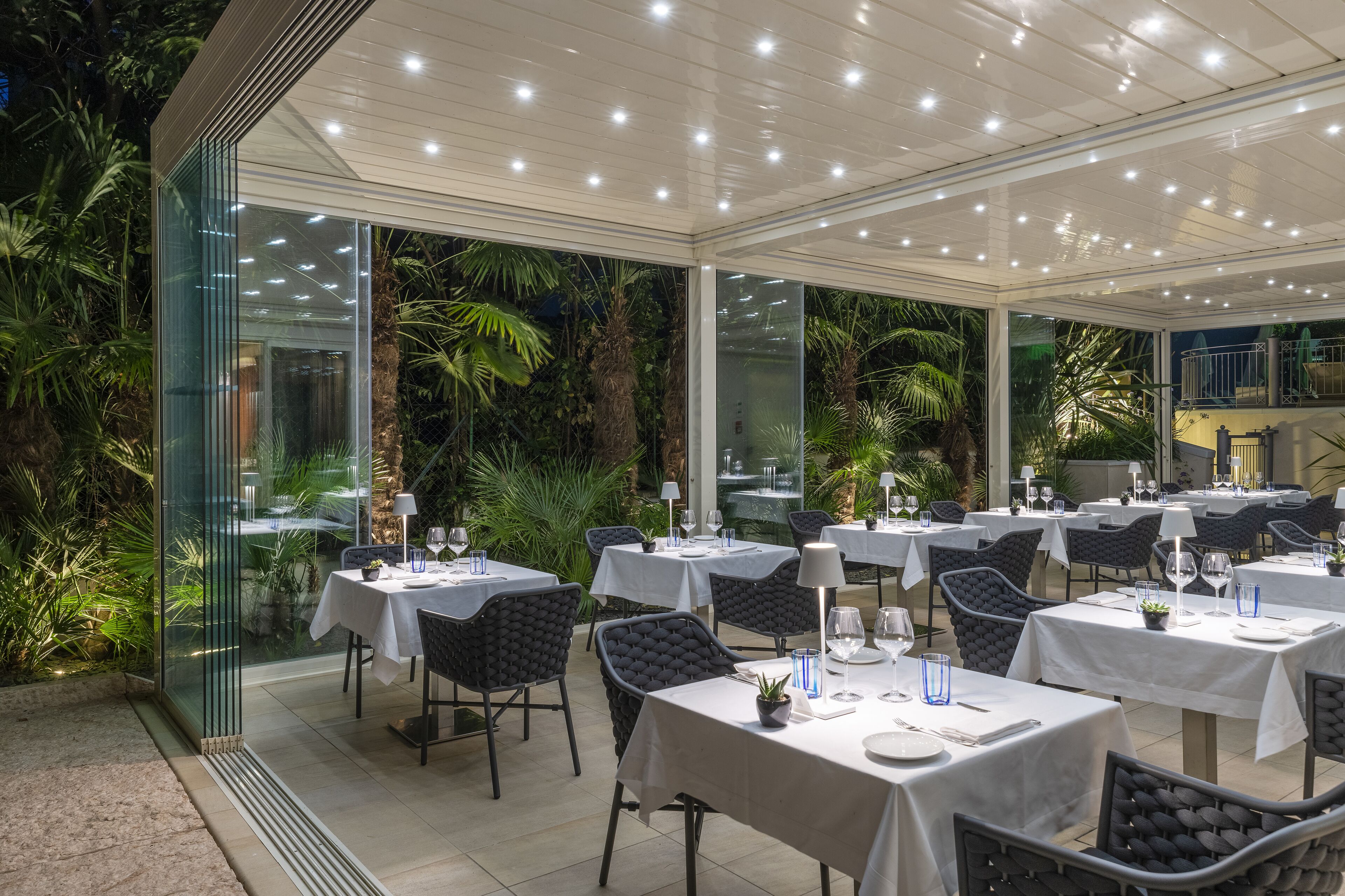 A restaurant with glass walls, white tables, and black chairs, surrounded by lush greenery.