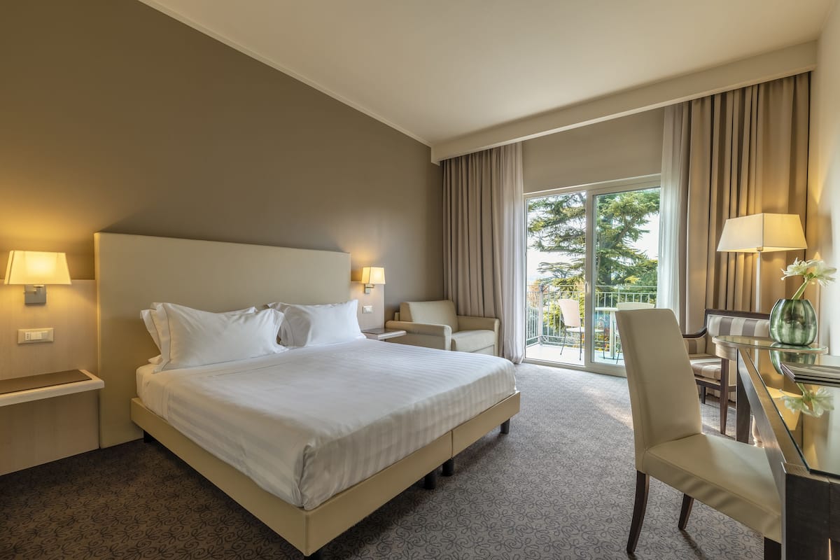 A hotel room with a large bed, two bedside tables with lamps, a desk with a chair, and a view of a balcony with curtains.