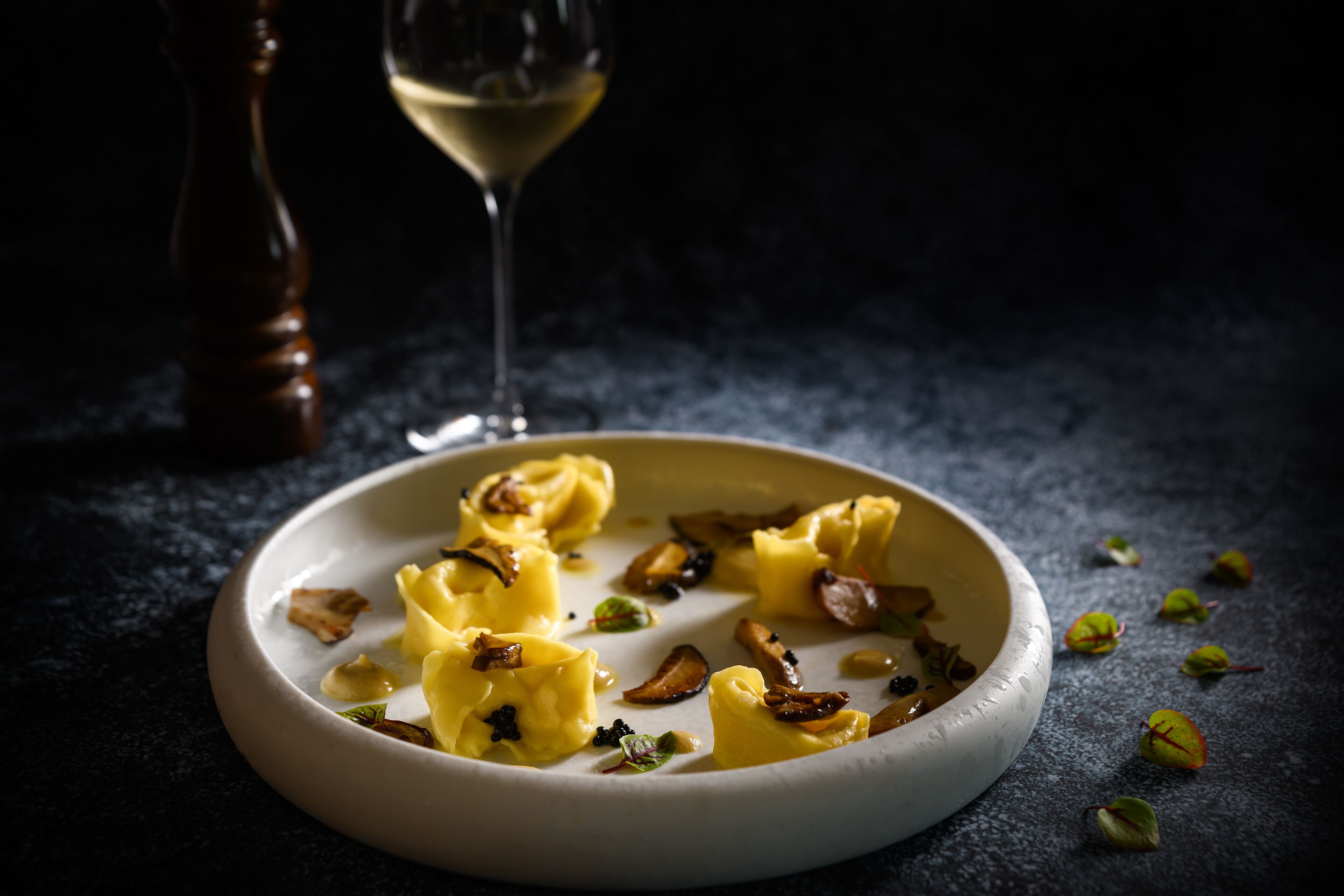 En tallrik med ravioli, svamp och örter, ett glas vitt vin och löv utspridda på en mörk yta.