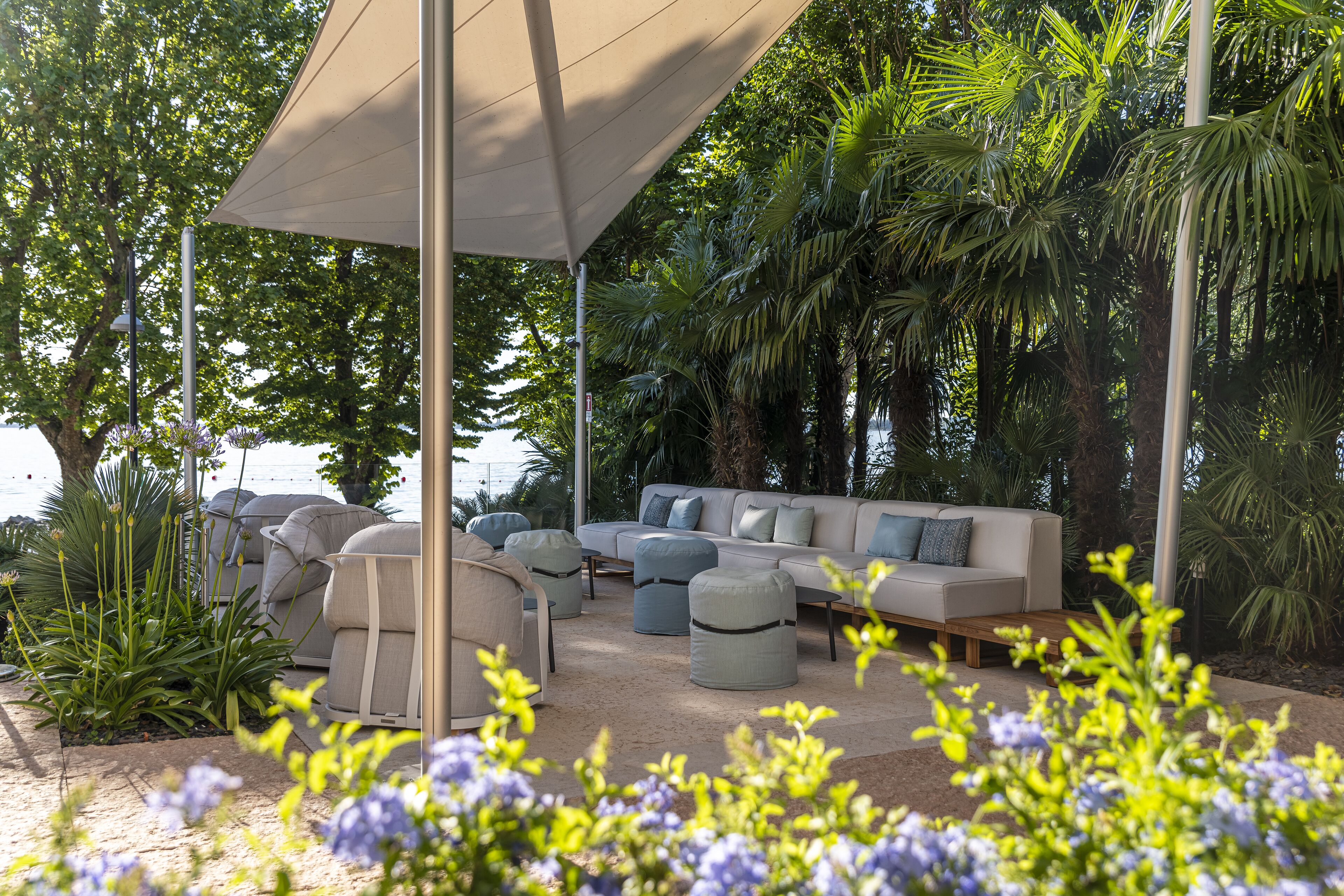 Un espace extérieur lumineux, aménagé avec un canapé et des poufs, entouré d’une végétation luxuriante et surmonté d’une tonnelle.