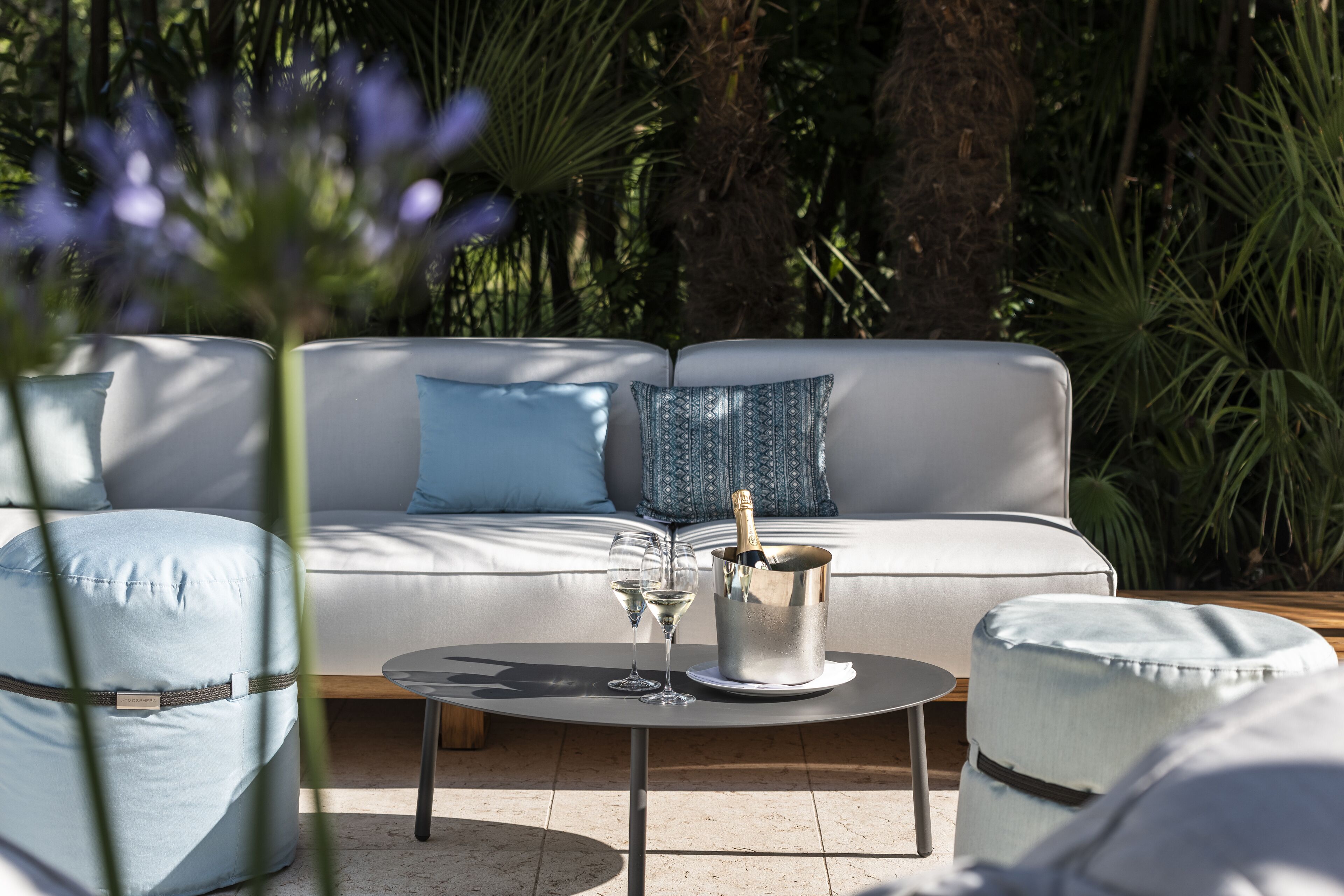 A modern outdoor seating area with a white sofa, two blue cushions, a round table with a champagne bottle and glasses, and a wooden floor.