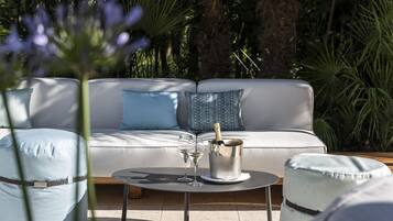 A modern outdoor seating area with a white sofa, two blue cushions, a round table with a champagne bottle and glasses, and a wooden floor.