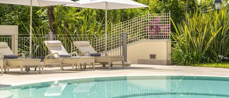 Zona de piscina con sillones de descanso y sombrillas, rodeada de vegetación exuberante y una valla decorativa.