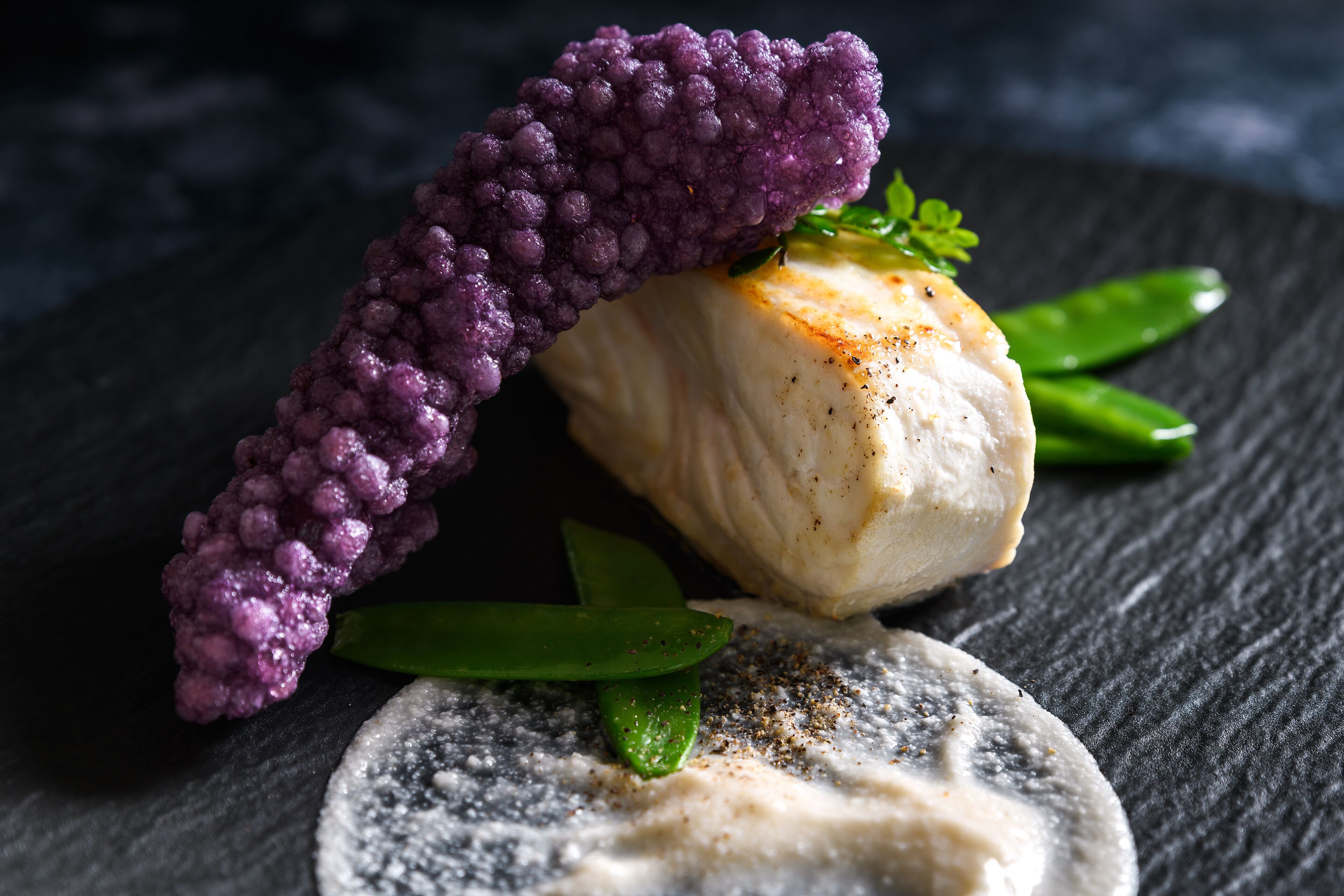 A dish with a piece of white fish, green asparagus, and purple garnish on a dark slate plate.