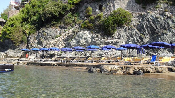 A coastal beach with clear water, a rocky cliff, and a row of sun loungers under blue umbrellas. - Hotel Ristorante Baia del Sorriso (Rosignano Marittimo)