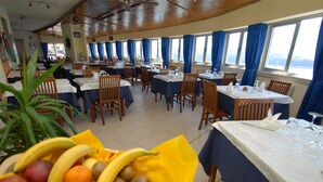 A dining area with tables set for meals, a fruit basket in the foreground, and large windows with curtains. - Hotel Ristorante Baia del Sorriso (Rosignano Marittimo)