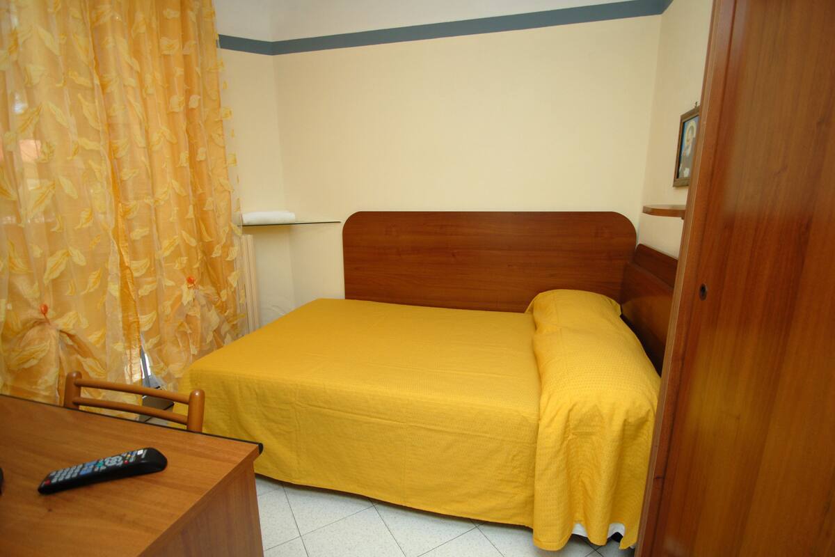A small, single-bed room with a wooden headboard, a yellow bedspread, a wooden desk with a remote control, and a chair.
