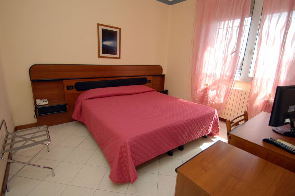 A bedroom with a wooden bed, a desk with a computer, a chair, and a window with curtains.