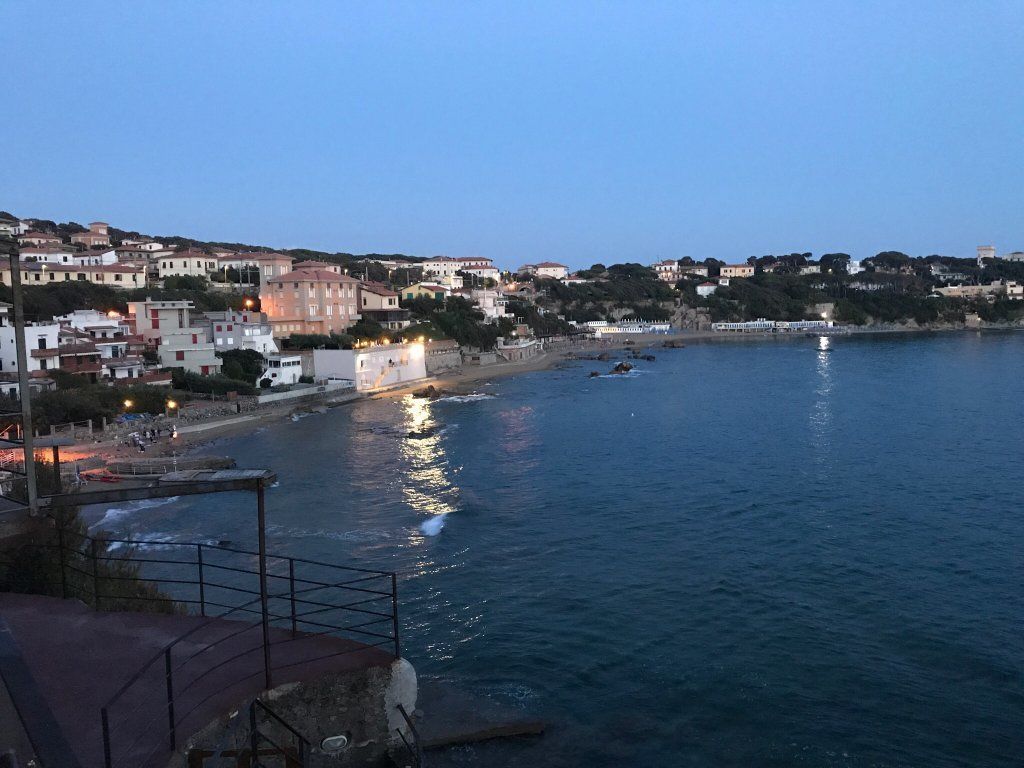 A coastal town at dusk with buildings along the waterfront, a pier, and boats in the water.
