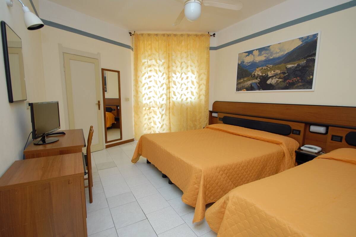 A hotel room with two beds, a desk, a chair, a mirror, a painting on the wall, and a ceiling fan.