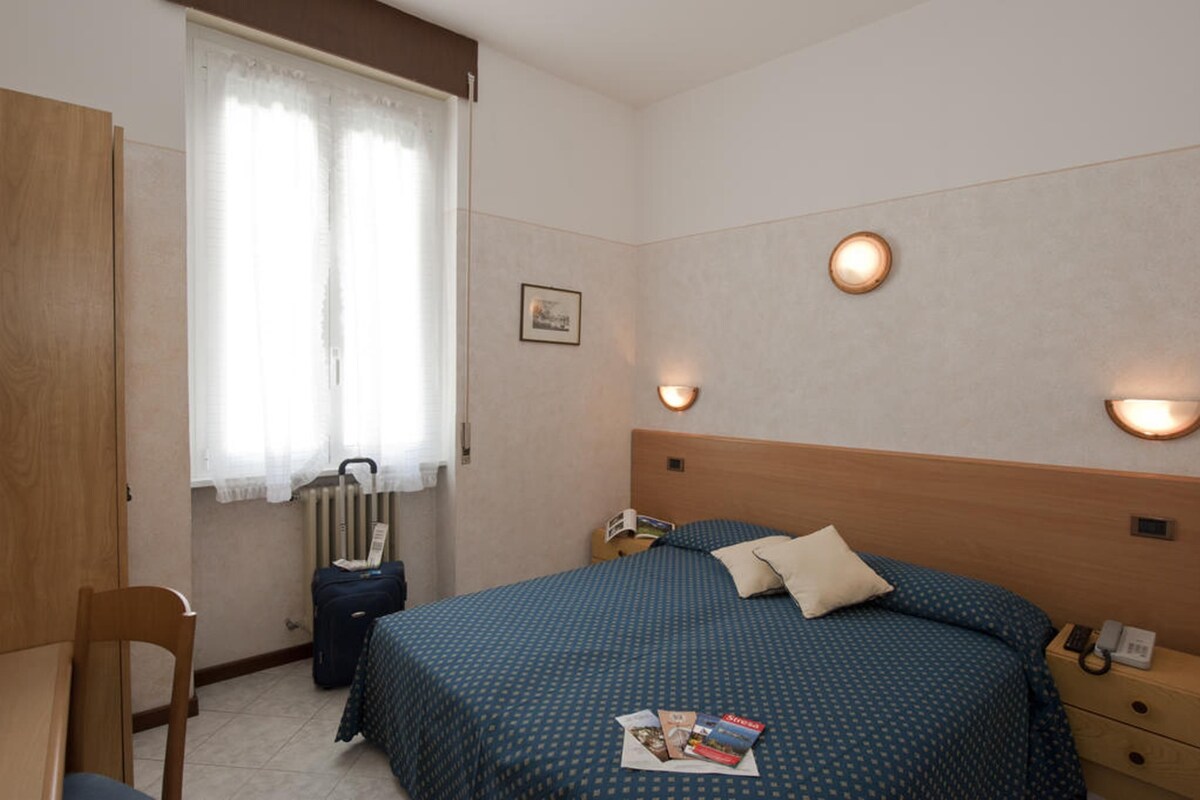 Une chambre d’hôtel avec un lit, des tables de chevet, une chaise, un radiateur et une fenêtre avec des rideaux.