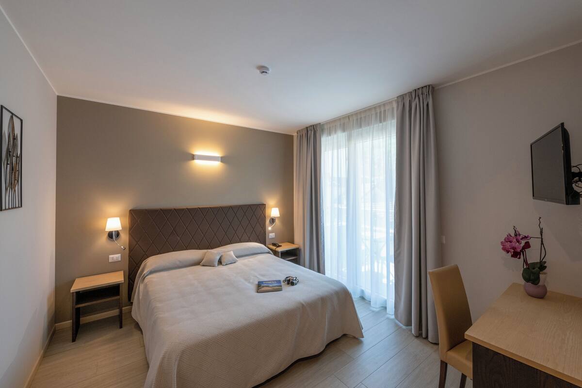 Habitación de hotel con una cama grande, lámparas de noche, una mesita, una silla y una planta en maceta.