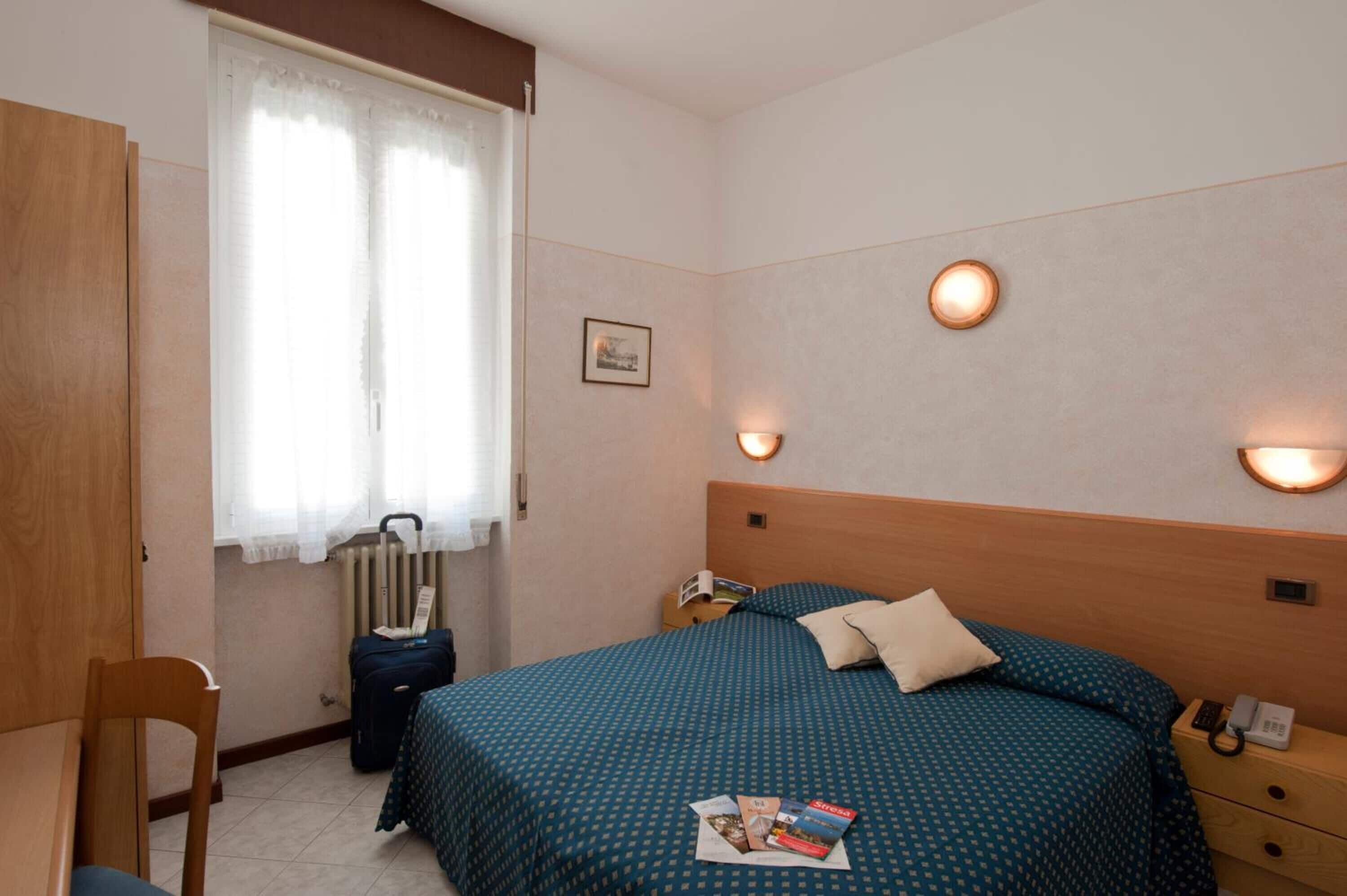 A hotel room with a bed, bedside lamps, a chair, a radiator, and a suitcase.