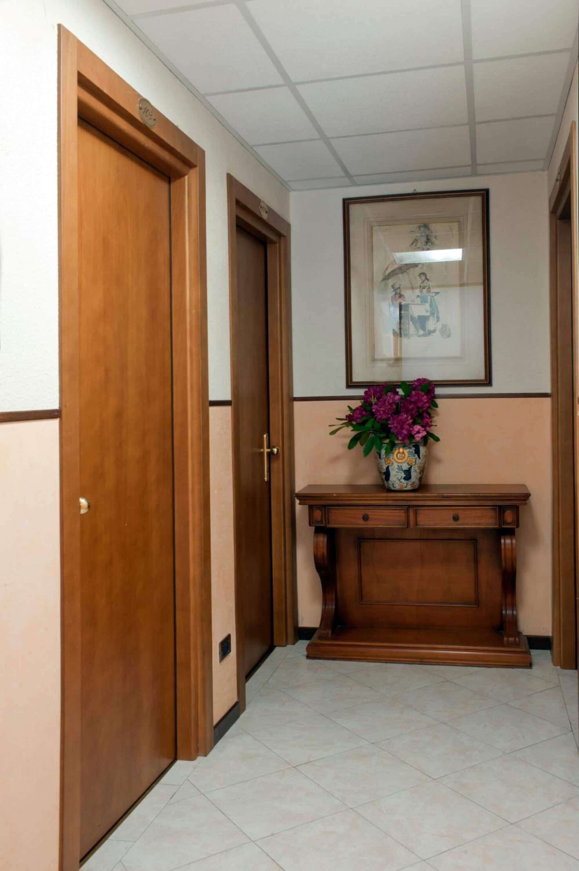 Un pasillo con puertas de madera, piso de baldosas, una consola de madera con un jarrón de flores y un cuadro en la pared.