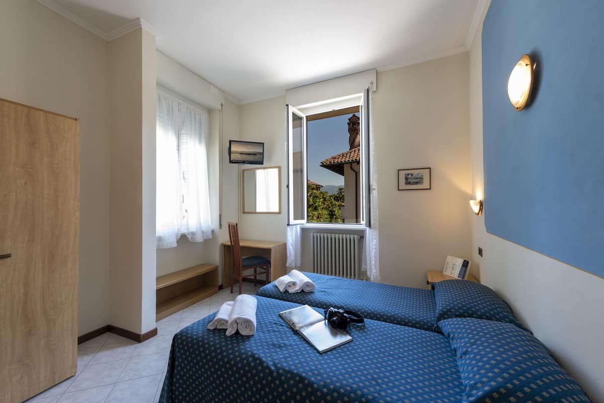 Habitación de hotel con dos camas, un escritorio, una silla y una ventana que ofrece vistas a un edificio y árboles.