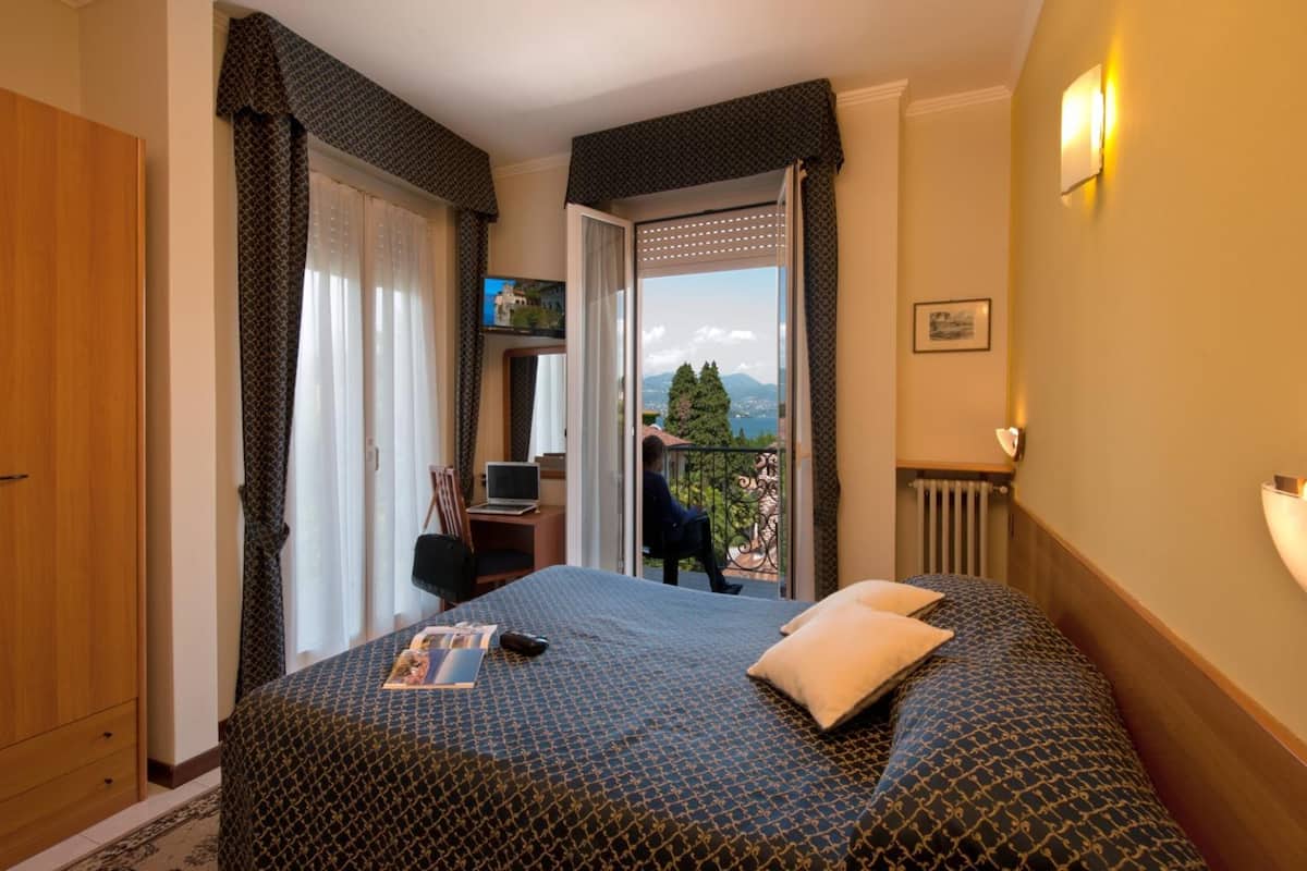 Habitación de hotel con cama, escritorio con computadora portátil, televisor y balcón con vistas a las montañas.
