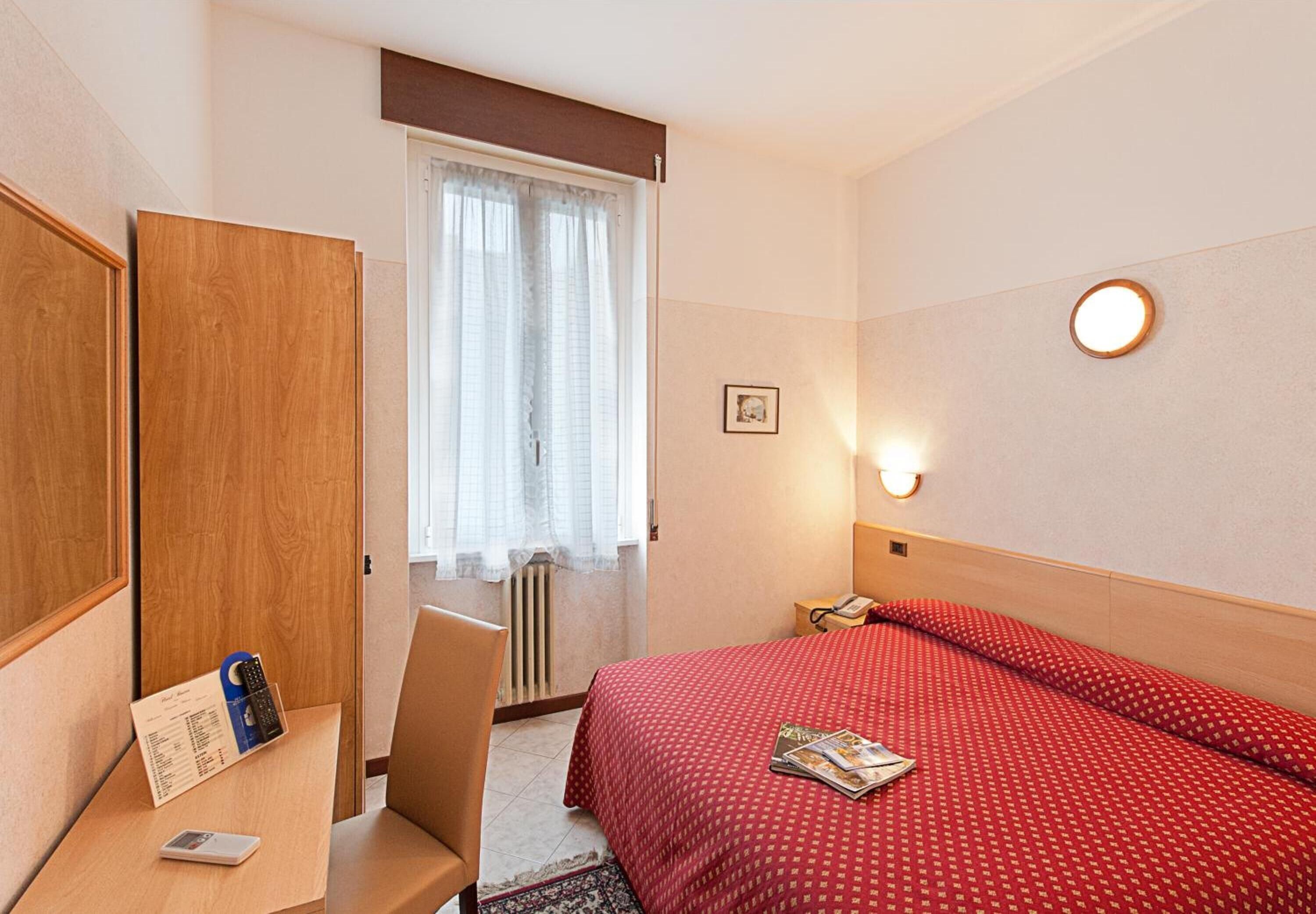 A hotel room with a bed, a desk with a telephone and a remote control, a chair, a window with curtains, and a wall-mounted light fixture.