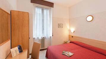 Hotelkamer met een bed, een bureau met telefoon en afstandsbediening, een stoel, een raam met gordijnen en een wandlamp.
