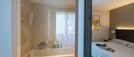 A modern hotel room with a glass-enclosed shower, a bed with a headboard, and a wall-mounted nightstand.