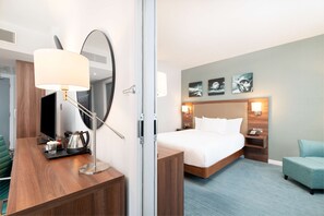 A hotel room with a bed, bedside tables, a desk with a lamp, a chair, and artwork on the wall. - Hilton Garden Inn London Heathrow Airport (Hounslow)
