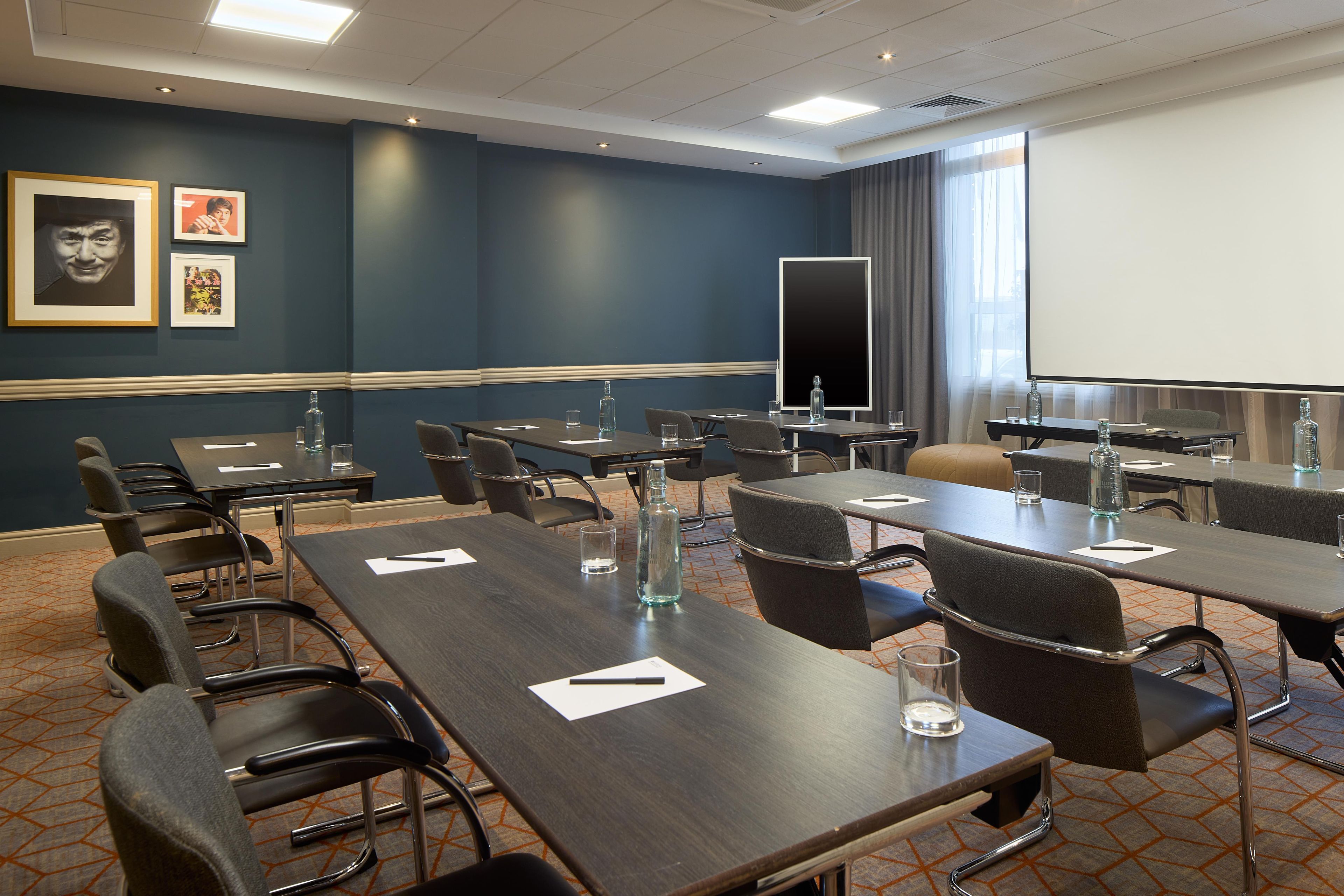 Ein Konferenzraum mit mehreren Tischen und Stühlen, einer Projektionsleinwand und gerahmten Kunstwerken an der Wand.