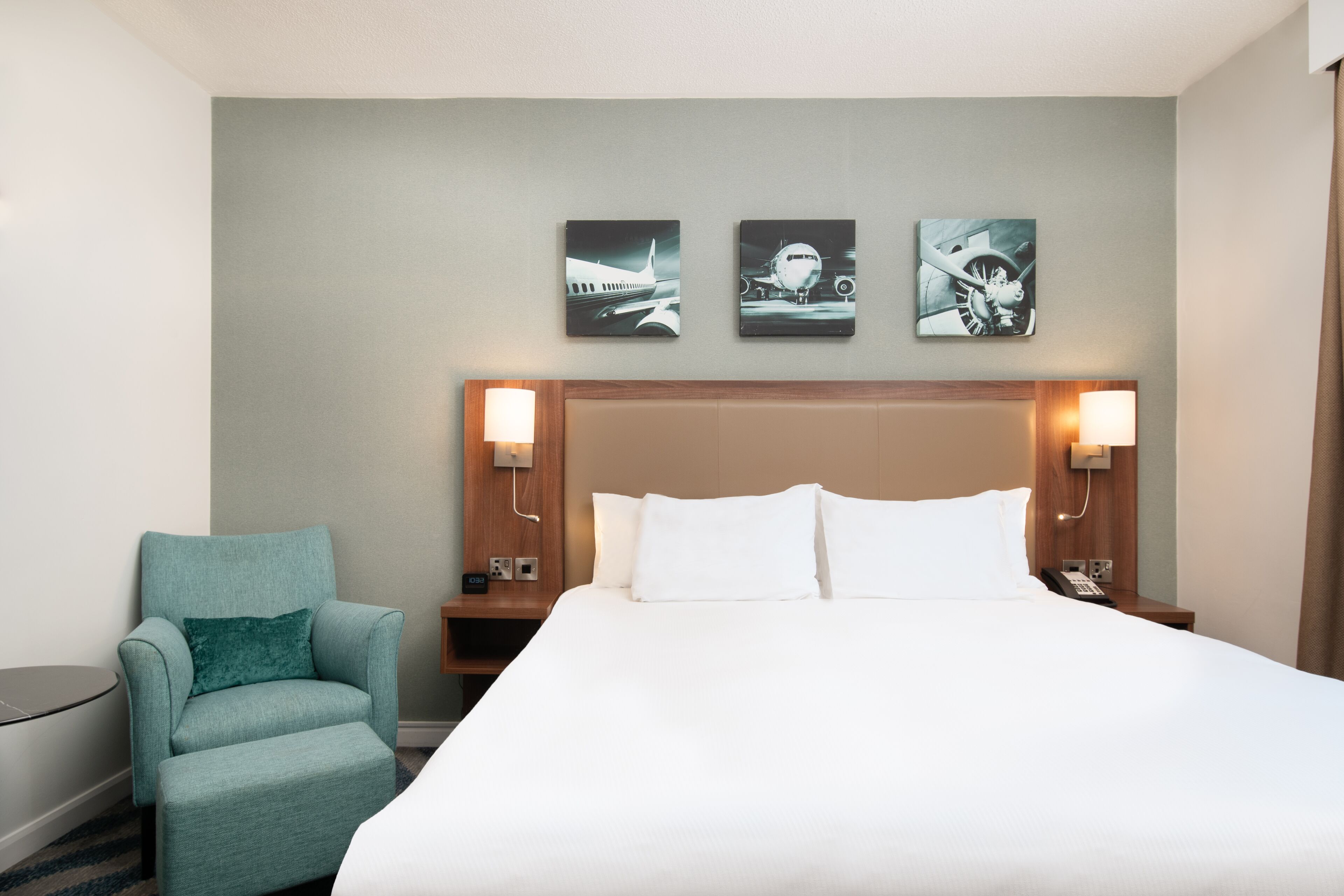 A hotel room with a large bed, a teal armchair, and three wall-mounted artworks depicting airplanes.