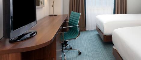 A hotel room with a flat-screen TV, a desk with a lamp, a green chair, a bed, and a window with curtains.