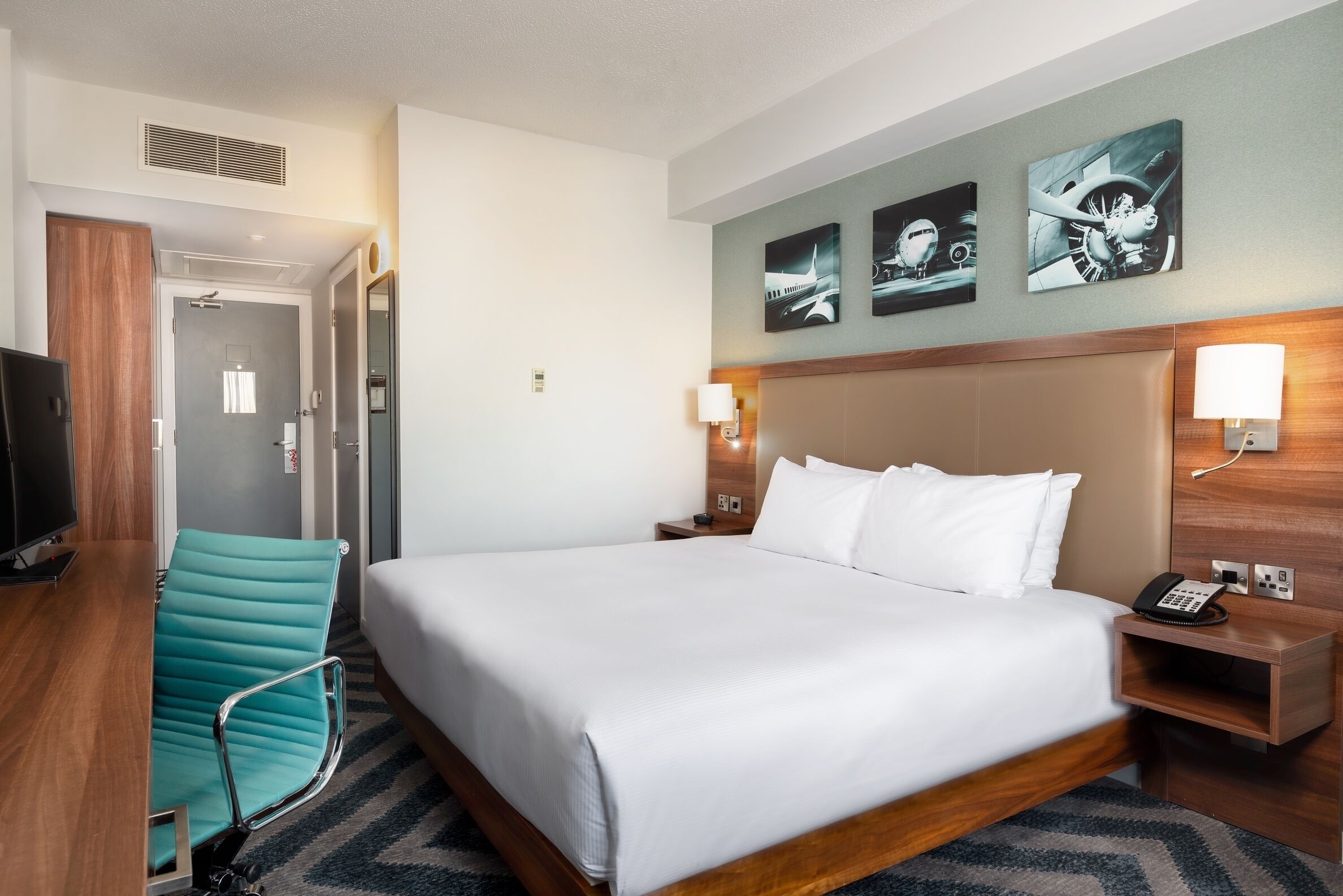 A hotel room with a large bed, a desk with a chair, a TV, and artwork on the wall.