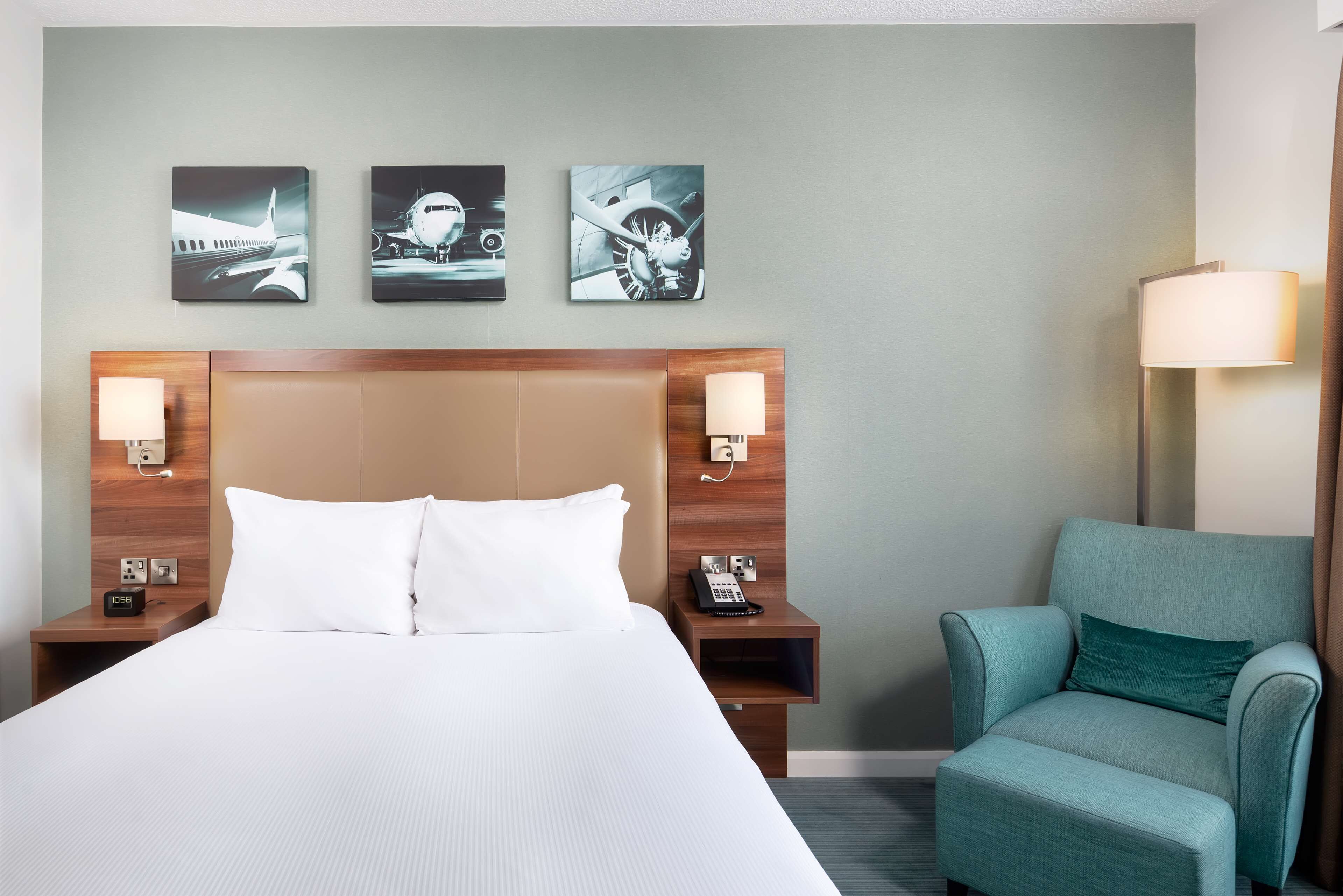 Une chambre d’hôtel avec un grand lit, deux tables de chevet avec des lampes, une chaise et trois œuvres d’art accrochées au mur, représentant des avions.