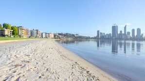 A beach with a calm water body and a city skyline in the background. - The Peninsula Riverside Serviced Apartments (South Perth)