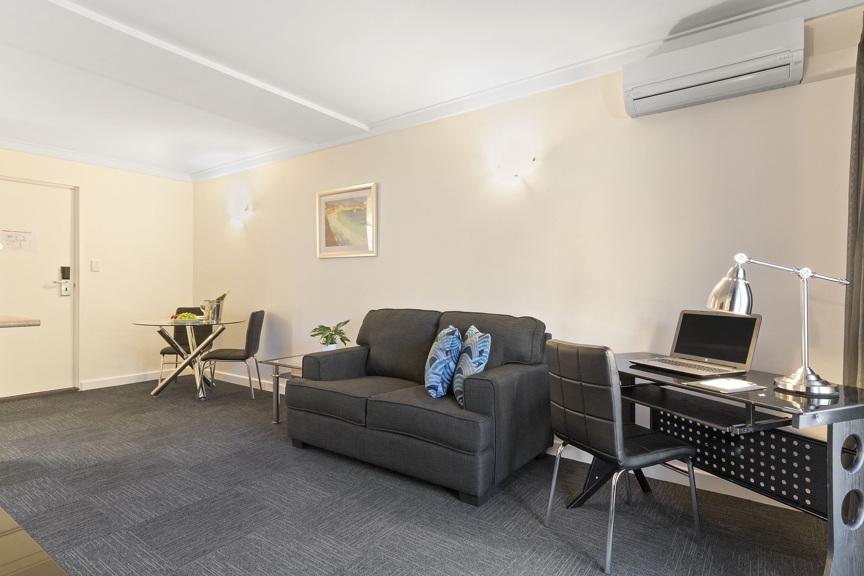 Een moderne hotelkamer met een grijze bank, een eettafeltje, een bureau met een laptop en een airconditioner aan de muur.