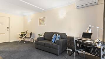 Een moderne hotelkamer met een grijze bank, een eettafeltje, een bureau met een laptop en een airconditioner aan de muur.