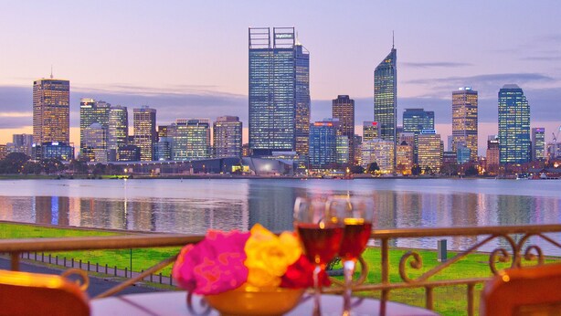 A table with a bowl of flowers and two glasses of red wine, overlooking a city skyline with high-rise buildings and a body of water. - The Peninsula Riverside Serviced Apartments (South Perth)