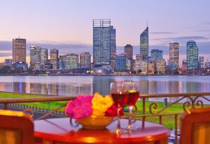 A table with a bowl of flowers and two glasses of red wine, overlooking a city skyline with high-rise buildings and a body of water. - The Peninsula Riverside Serviced Apartments (South Perth)