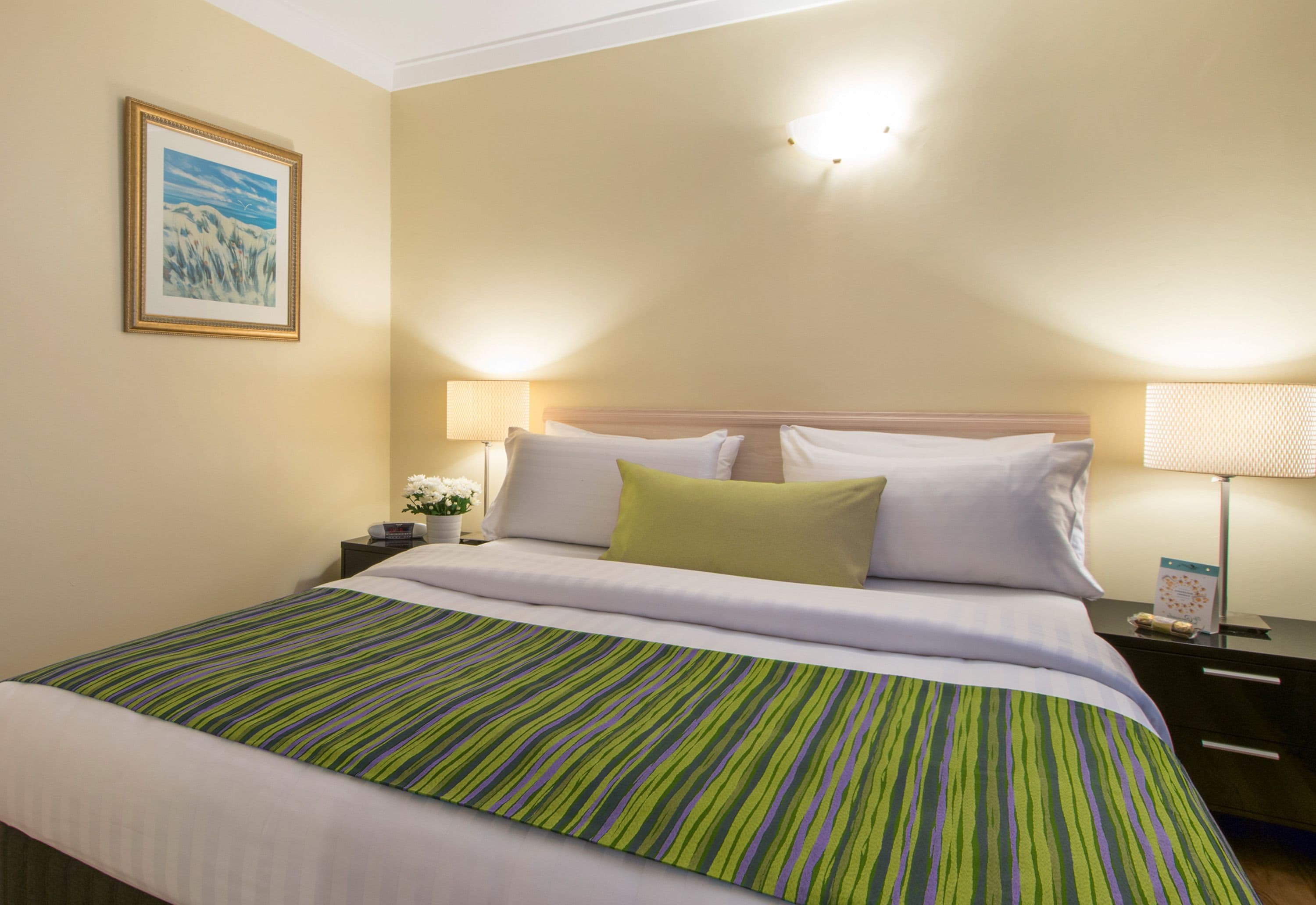 Habitación de hotel con cama, dos mesitas de noche, lámpara, un cuadro en la pared y ventana con persianas.