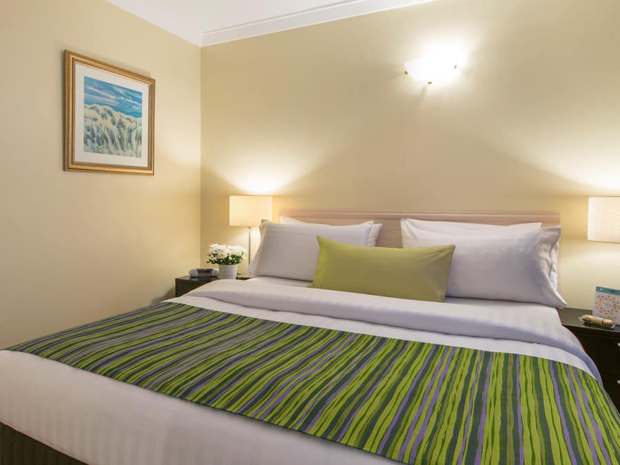 Habitación de hotel con cama, dos mesitas de noche, lámpara, un cuadro en la pared y ventana con persianas.