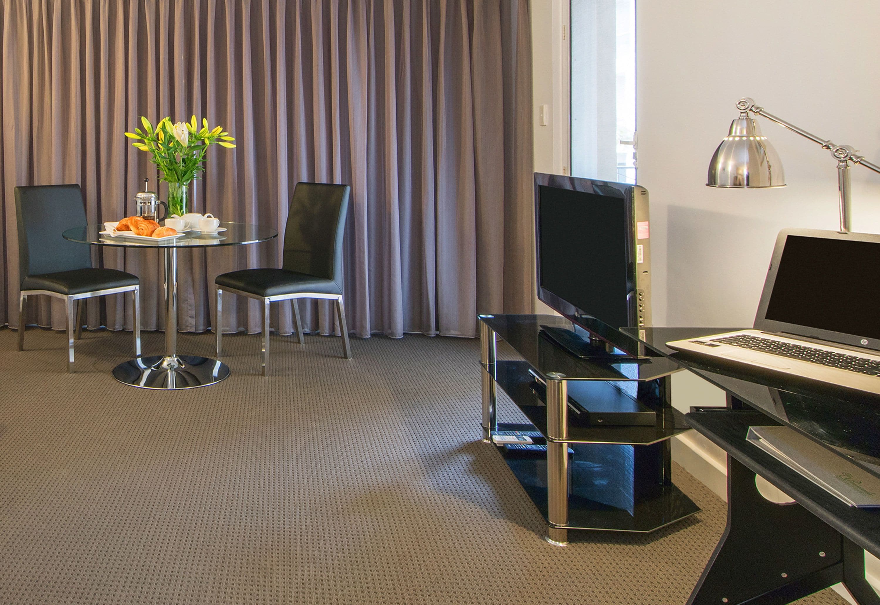 Habitación de hotel con una mesa de vidrio, dos sillas, un televisor, una computadora portátil, una lámpara y un jarrón con flores.