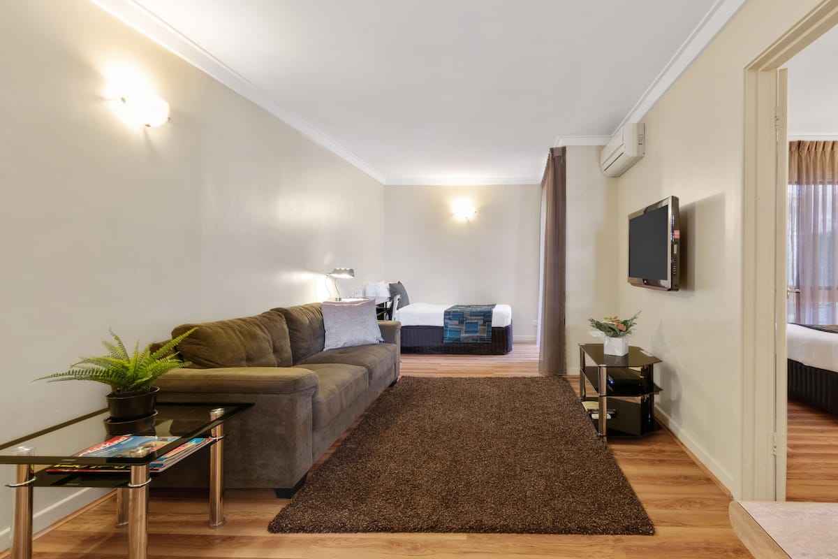 A modern living room with a sofa, a coffee table, a TV mounted on the wall, and a bed in the background.