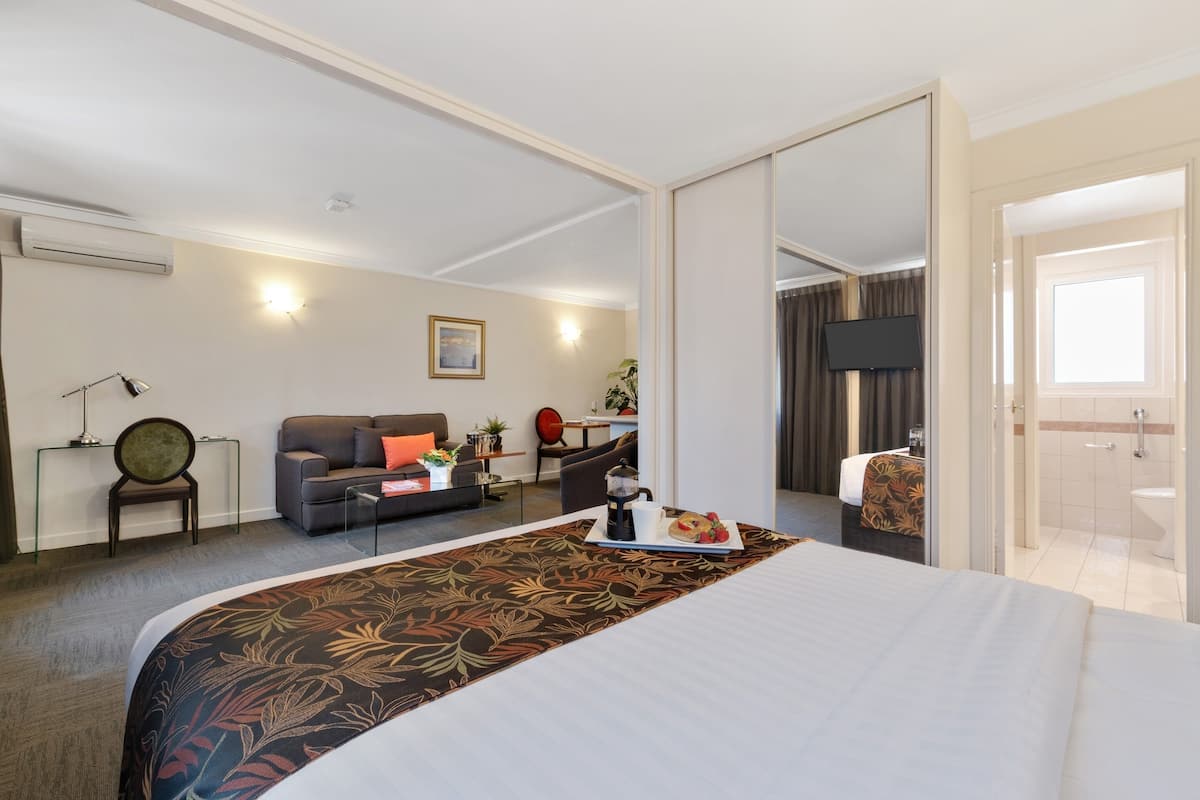 A hotel room with a large bed, a sofa, a coffee table, and a desk. There is a bathroom visible through an open door.