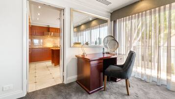 Habitación con escritorio, silla y un espejo grande. Se ve una ventana con cortinas traslúcidas y vistas al exterior con zonas verdes.