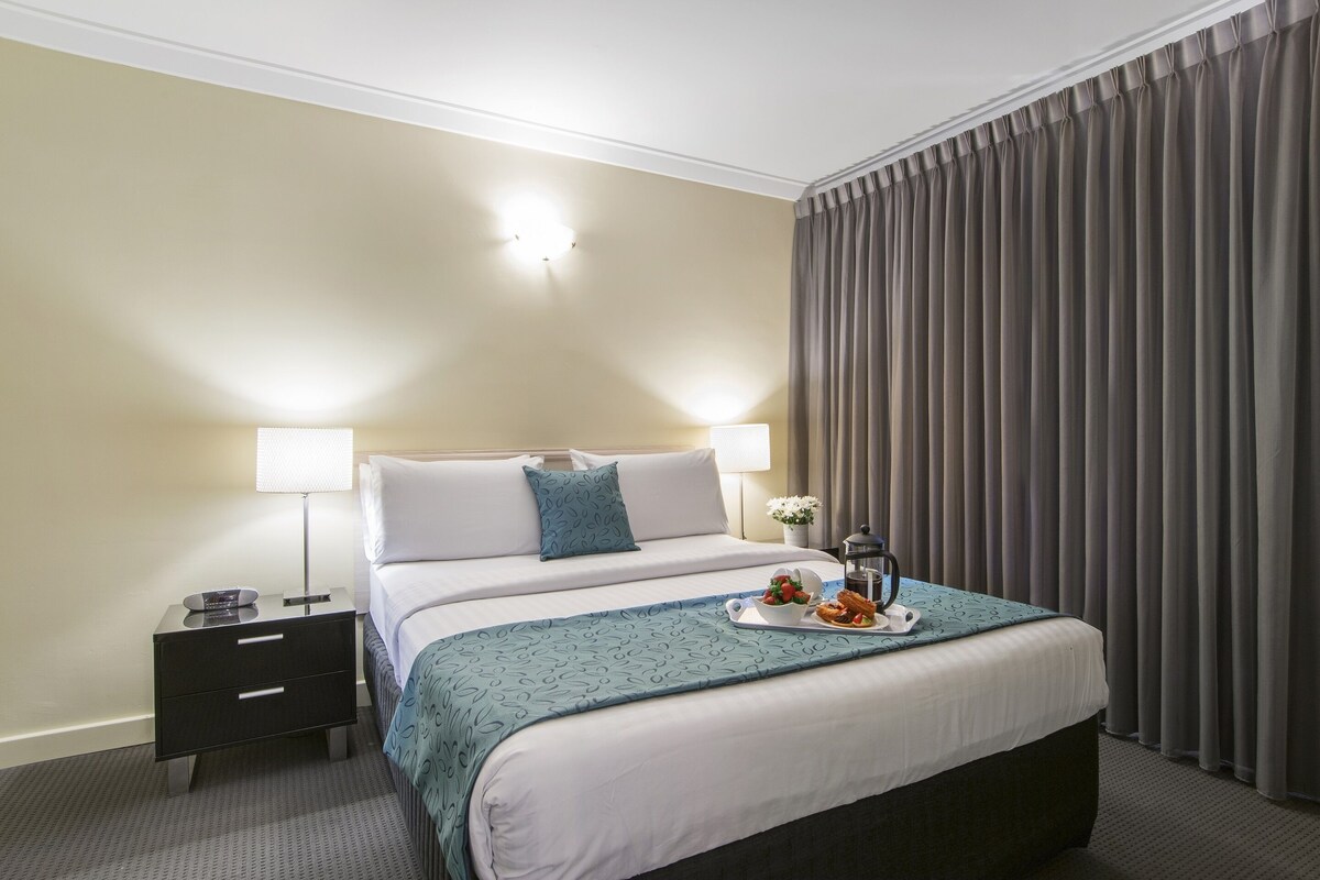 A hotel room with a bed, bedside lamps, a nightstand, a tray with food, and a window with curtains.