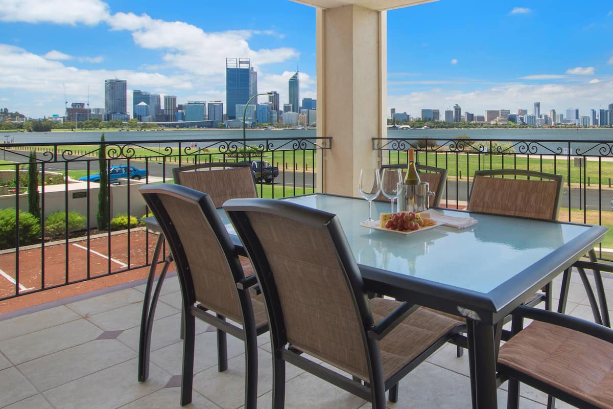 A balcony with a glass table and chairs, overlooking a city skyline and a body of water.