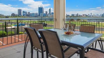 Uma varanda com mesa e cadeiras de vidro, com vista para o horizonte urbano e um corpo de água.