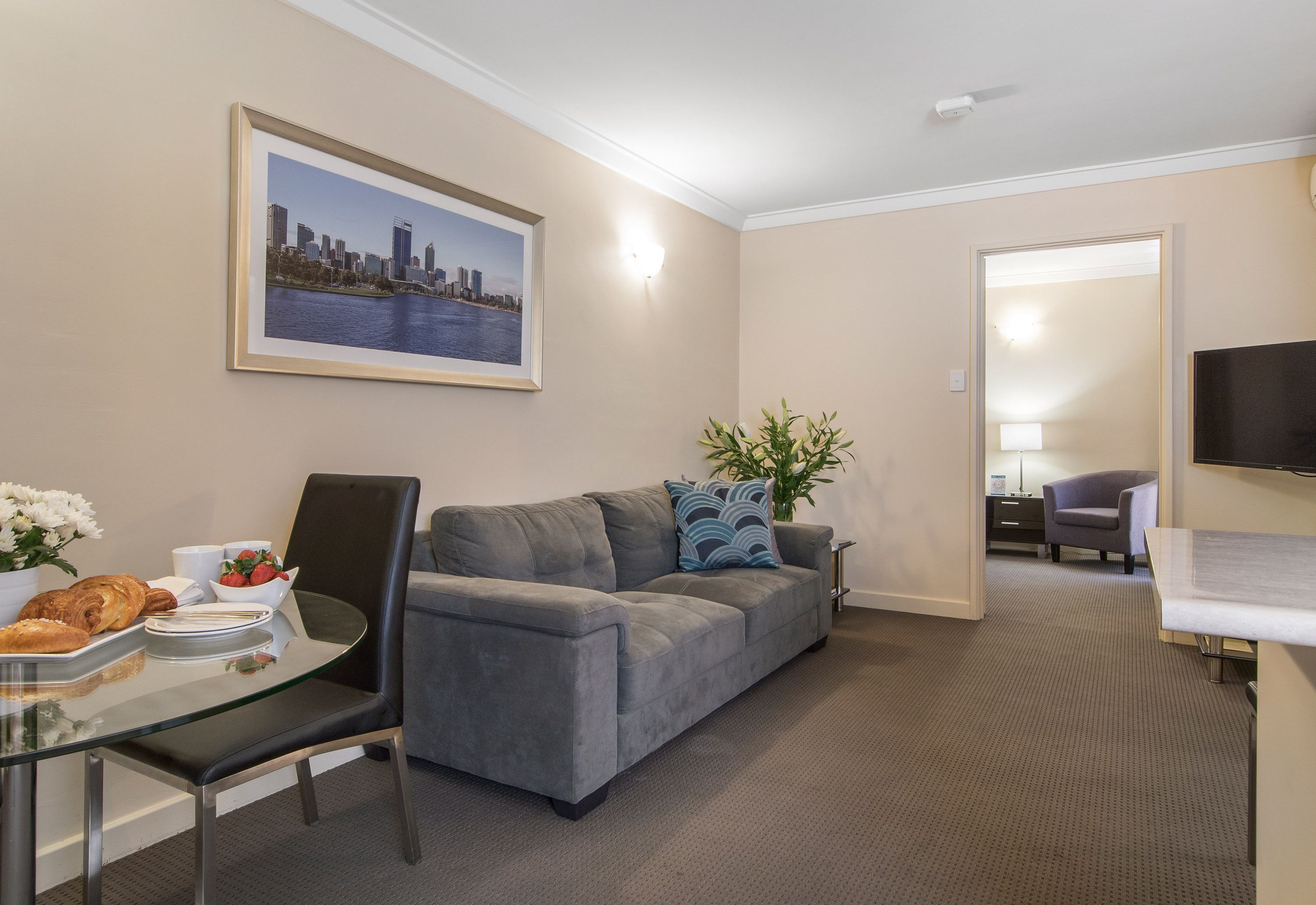 Un hotel moderno con un sofá gris, una mesa de comedor con alimentos para el desayuno, un cuadro con una vista urbana enmarcada y un televisor en la pared.