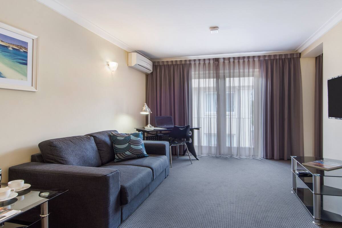 A modern hotel room with a grey sofa, a desk with a laptop, a TV, and a large window with curtains.