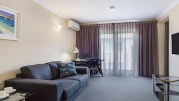 Una habitación de hotel moderna con un sofá gris, un escritorio con una computadora portátil, un televisor y un amplio ventanal con cortinas.