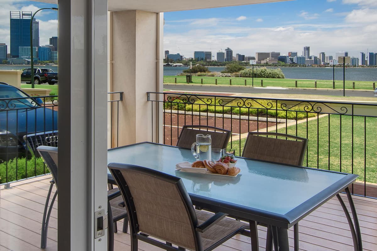 A balcony with a glass table, chairs, and a tray of pastries. A city skyline and a body of water are visible in the background.