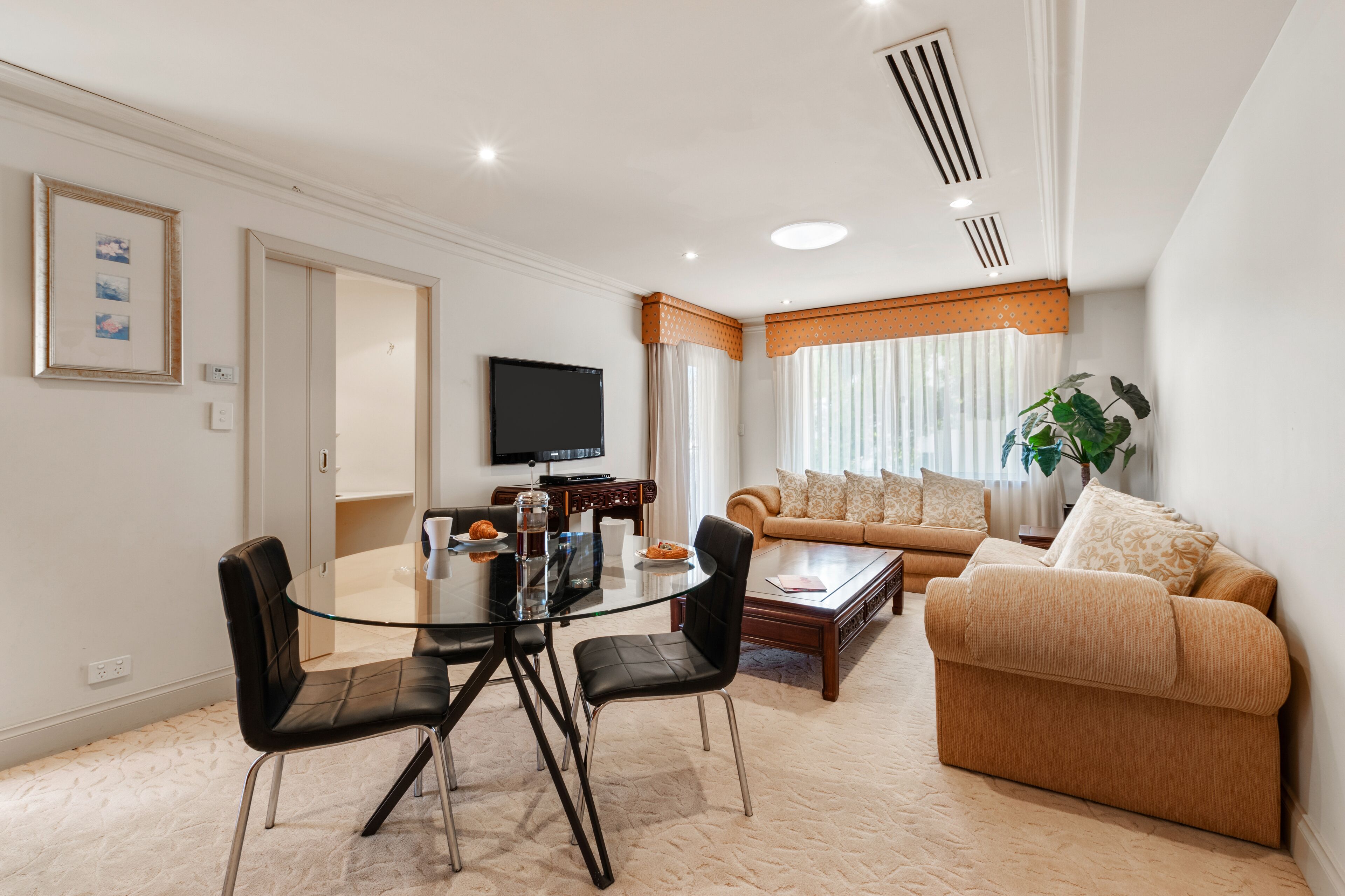 A modern living room with a glass dining table, black chairs, a beige sofa, and a television on the wall.
