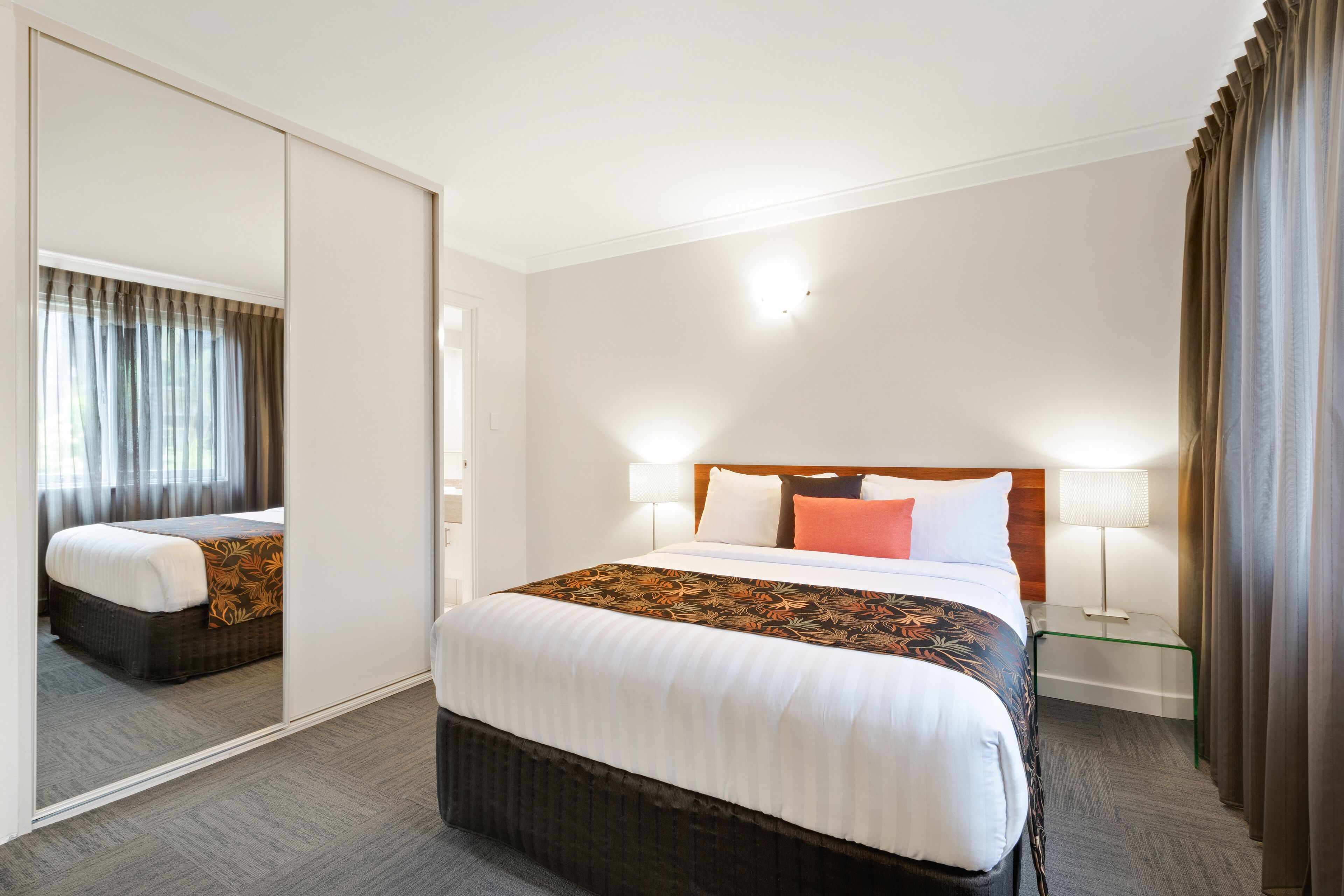 A hotel room with a large bed, a mirror, a bedside table with a lamp, and a view of the outside through a window with curtains.