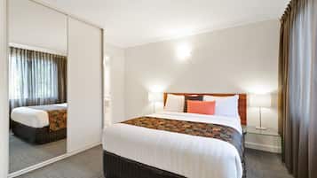 A hotel room with a large bed, a mirror, a bedside table with a lamp, and a view of the outside through a window with curtains.